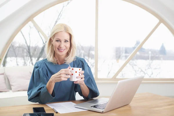Smiling sales businesswoman having coffee while working at her office desk on the laptop.
