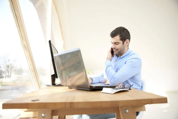 彼の携帯電話で誰かと話して 机に座っている間のラップトップに取り組んでカジュアルな青年実業家 ホーム オフィス — ストック写真