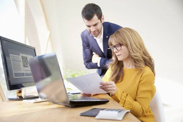 Middle aged businesswoman sitting at office desk and holding a document in her hands while young financial assistant businessman standing next to her and consulting about the new project. Teamwork at the office.