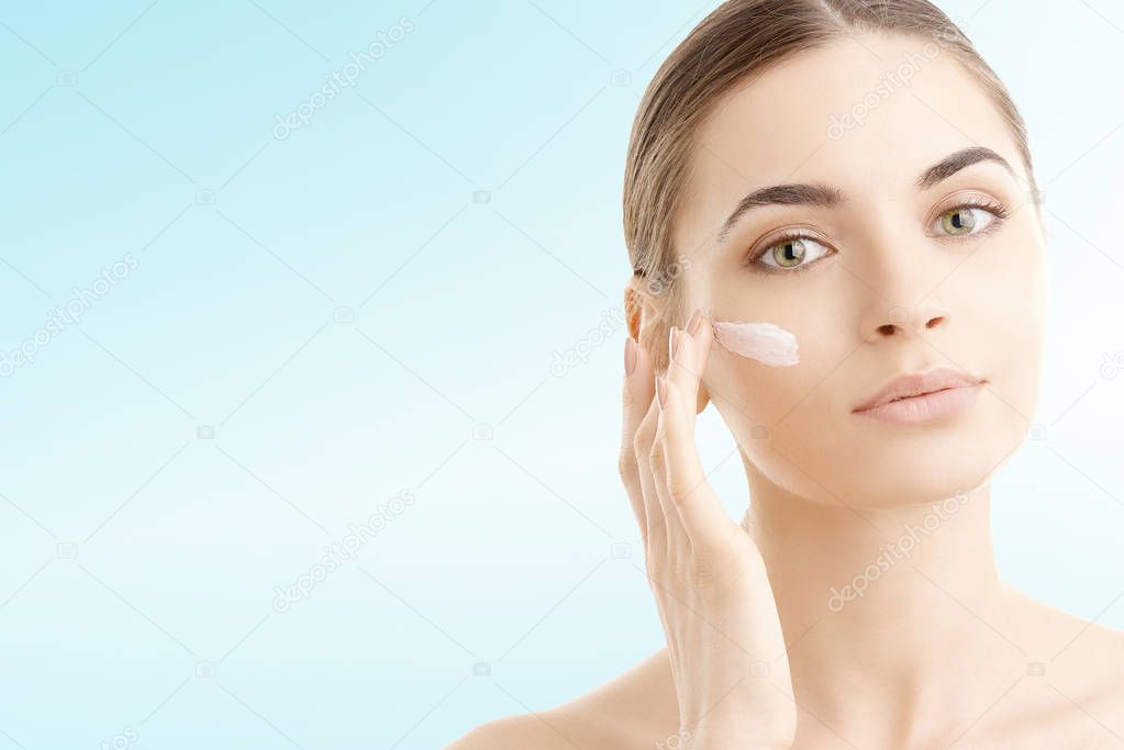Studio shot of a beautiful young woman applying moisturizer onto her face against a blue background with copy space. 