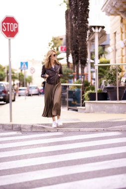 Orta yaşlı mutlu bir kadının yaya geçidinde yürüdüğü uzun bir çekim. Güneş gözlüğü takan kendine güvenen ve etekli motorcu ceketi giyen bir kadın.. 