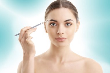 Güzel bir genç kadının onun kaş duran izole açık mavi renkli, makyaj uygulamak Studio vurdu. 