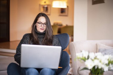 Sırıtkan genç bir kadının dizüstü bilgisayarı ile sandalyede otururken ve evde çalışırken çekilmiş fotoğrafı..