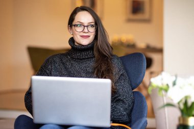 Sırıtkan genç bir kadının dizüstü bilgisayarı ile sandalyede otururken ve evde çalışırken çekilmiş fotoğrafı..