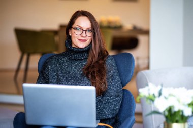 Sırıtkan genç bir kadının dizüstü bilgisayarı ile sandalyede otururken ve evde çalışırken çekilmiş fotoğrafı..