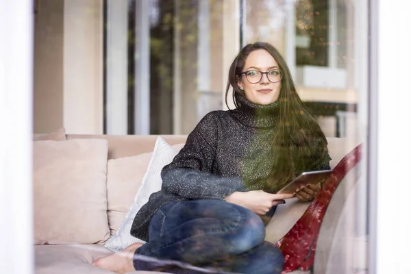 家でアームチェアでリラックスしながら デジタルタブレット上でネットサーフィンガラス窓の後ろに笑みを浮かべて女性のショット — ストック写真