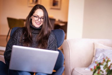 Koltukta otururken ve evde çalışırken gülümseyen bir kadının not defteri kullanması. Merkez Ofis.