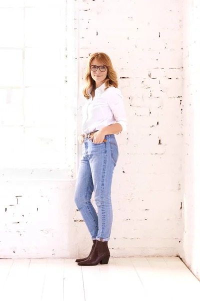 中年妇女穿着白衬衫和蓝色牛仔裤在白砖墙边放松时的全照 — 图库照片