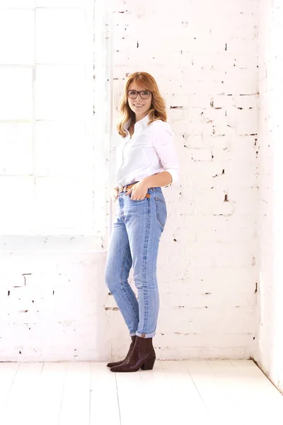 中年妇女穿着白衬衫和蓝色牛仔裤在白砖墙边放松时的全照 — 图库照片