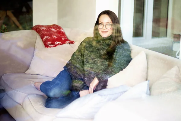 家で快適なソファでリラックスしながら タートルネックのセーターとジーンズを身に着けている窓の後ろに笑顔の若い女性のショット — ストック写真