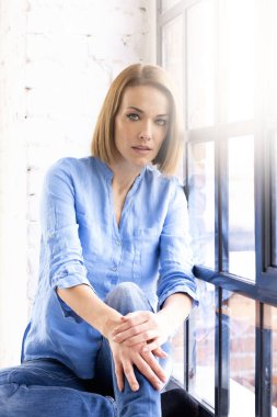 Pencere kenarında dinlenirken gömlek ve kot giyen çekici orta yaşlı kadının portresi..