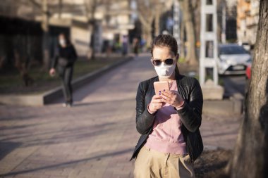 Coronavirus salgını sırasında solunum maskesi takan genç bir kadının fotoğrafı. Kendine güvenen bir kadın sokakta bankta otururken mesaj atıyor.. 