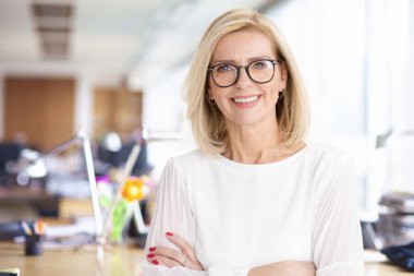 Ofiste kollarını kavuşturmuş, kameraya bakıp gülümseyen çekici bir iş kadınının portresi..