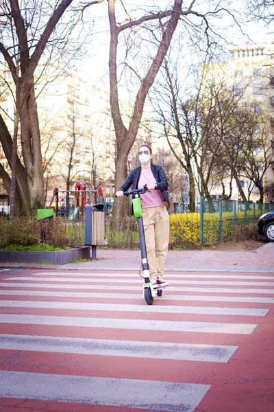 街で電動スクーターに乗っている間顔マスクを身に着けている若い女性の完全な長さのショット — ストック写真