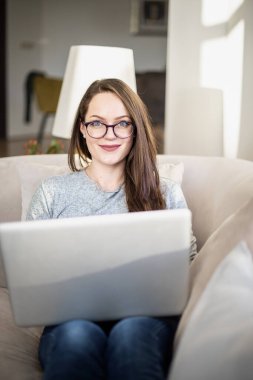 Evdeki kanepede otururken dizüstü bilgisayar kullanan gülümseyen genç bir kadının yakın çekimi. Merkez Ofis. 