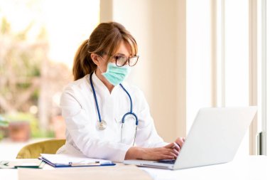 Masada otururken ve hastanede dizüstü bilgisayar üzerinde çalışırken yüz maskesi takan çok çalışan kadın doktor..