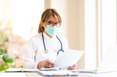Masada otururken ve hastanede dizüstü bilgisayar üzerinde çalışırken yüz maskesi takan çok çalışan kadın doktor..