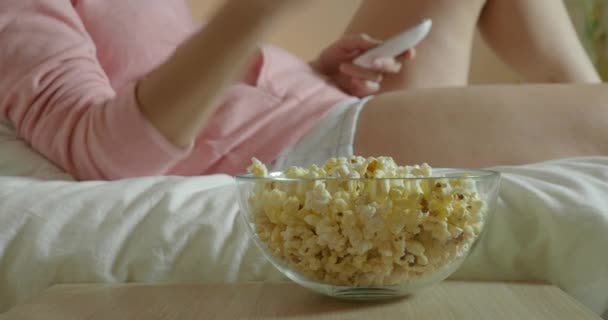 Женщина смотрит телевизор в спальне, лежит на кровати и ест попкорн — стоковое видео