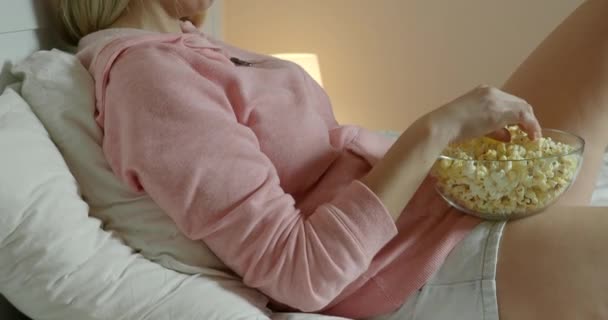 Mujer viendo la televisión en el dormitorio acostada en la cama y comiendo palomitas de maíz — Vídeo de stock