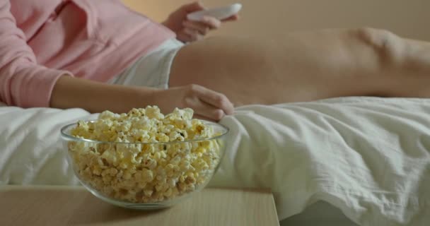 Mujer viendo la televisión en el dormitorio acostada en la cama y comiendo palomitas de maíz — Vídeos de Stock