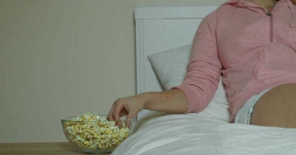 Женщина смотрит фильм лежит в постели и ест попкорн — стоковое видео