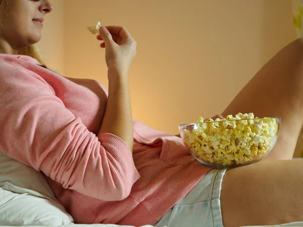 Женщина смотрит телевизор в спальне, лежит на кровати и ест попкорн — стоковое фото