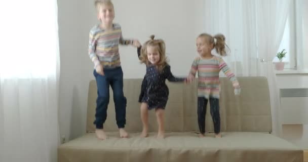 Två lillasyster och bror skrattar och hoppar på soffan — Stockvideo