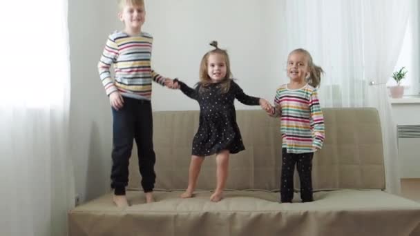 Две младшие сестры и брат смеются и прыгают на диване, замедленная съемка — стоковое видео