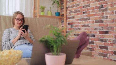 Oturma odasında kanepede oturan ve akıllı telefondan alışveriş yapan genç bir kadın.