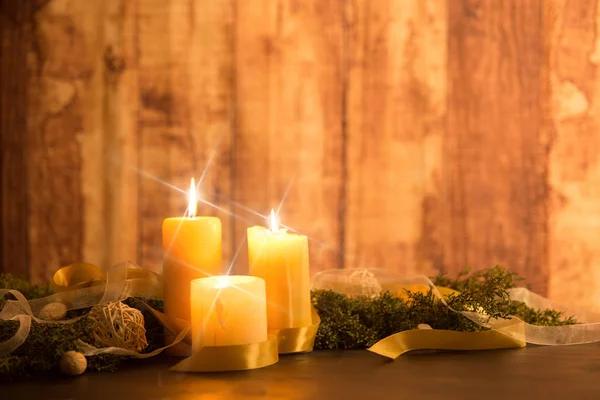 Тепло рождественской концепции: три свечи, зажженные крестовыми звездами, воздействуют на темный деревянный стол с сосновыми ветвями, натуральные сосновые шишки с золотым атласом и белые органзовые ленты — стоковое фото