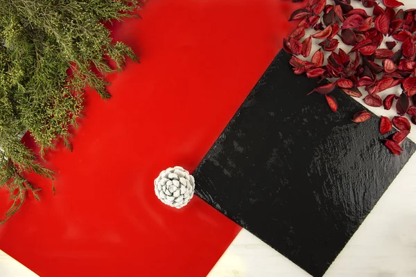 Flach liegender geometrischer Weihnachtskopierraum mit diagonalem roten Streifen, schwarzem Kalksteinteller, Tannenzweigen, weißem Tannenzapfen und roten Blütenblättern auf hellem Holzgrund — Stockfoto