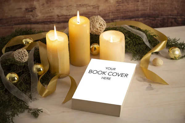Bir kitap tanıtımı için Noel ayini: boş kapaklı kitap üç yanan mum, beyaz organza ve altın saten kurdeleler, açık ahşap masa üzerinde altın mücevherler ve koyu ahşap arka plan ile yerleşmiş