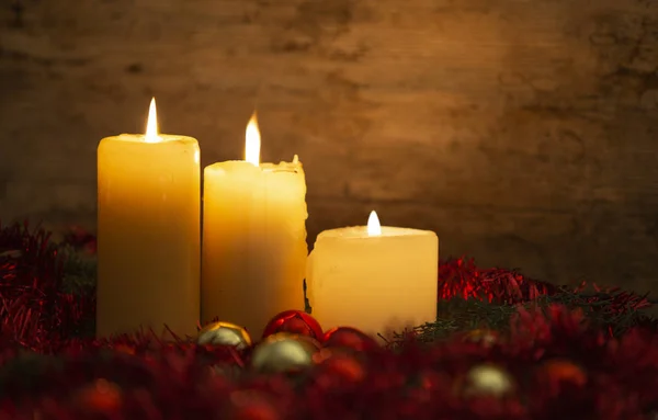 Тепло рождественской концепции: три свечи зажжены на светлом деревянном столе и деревенская обстановка с сосновыми ветвями, красное украшение и золотые и красные яркие безделушки с эффектом боке — стоковое фото