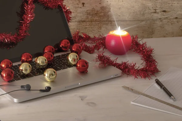 Trabajo de computadora en el concepto de vacaciones de Navidad: una computadora portátil de aluminio abierta, decoración de la corona roja, bolas rojas y doradas, vela encendida, pluma y regla en notas de bloque en la mesa de madera ligera — Foto de Stock