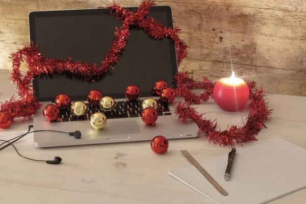 Trabajo de computadora en el concepto de vacaciones de Navidad: una computadora portátil de aluminio abierta, decoración de la corona roja, bolas rojas y doradas, vela encendida, pluma y regla en notas de bloque en la mesa de madera ligera — Foto de Stock