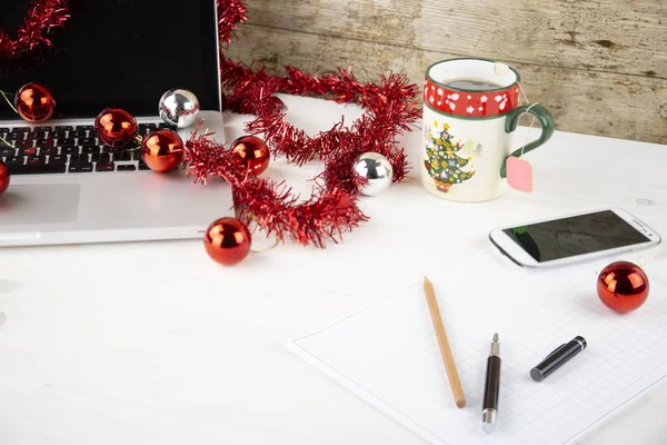 Computer job at Christmas holidays concept: light wooden table with an open aluminum laptop, red decoration, red baubles, Christmas cup of tea, smartphone, block notes, pencil and pen on block note