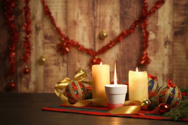Noel arkaplanında büyük alevli mum, çam dalları, dekupaj takılar, yanan mumlar ve ahşap arka planda asılı Noel süslemeleri ile birlikte.