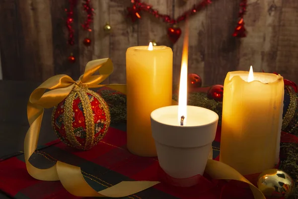 Крупный план зажженной свечи с большим пламенем на рождественской скатерти со спинными ветвями, декупаж безделушек, с зажженными свечами и вешанием рождественского украшения на деревянном фоне с эффектом боке — стоковое фото