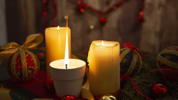 Крупный план зажженной свечи с большим пламенем на рождественской скатерти со спинными ветвями, декупаж безделушек, с зажженными свечами и вешанием рождественского украшения на деревянном фоне с эффектом боке — стоковое фото