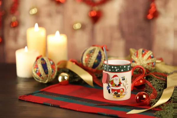 Taza de Navidad con etiqueta de té vacía en mantel de Navidad con alrededor de ramas de pino, adornos de decoupage, con velas encendidas y decoración de Navidad colgante sobre fondo de madera con efecto bokeh — Foto de Stock