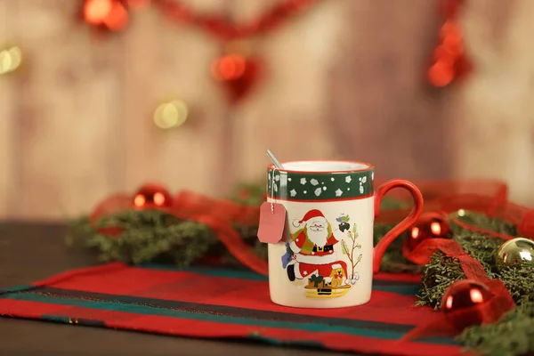 Noel bardağına boş çay etiketi asılmış, çam ağacı dalları, kırmızı mücevherler ve Noel süslemeleri bokeh efektli ahşap arka planda asılı.