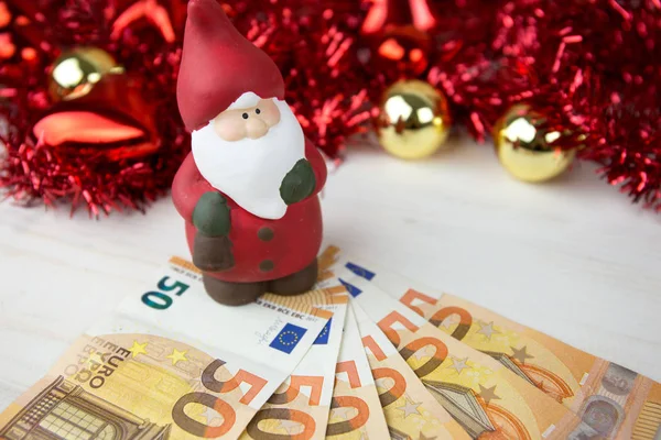 Noel para işi konsepti: Noel Baba heykelciği yaklaşık 50 avroluk banknotlar üzerinde kırmızı ve altın takılar ve bokeh etkisi olan çelenk süslemesi açık ahşap masa üzerinde