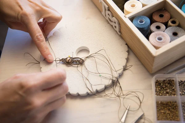 Lifestyle concept, work from home to reinvent your life: close-up of woman hands making macrame knotted jewelry with stone beads and tools on light wooden table — Stock Photo, Image