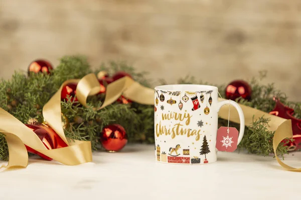 Primer plano de una taza de Navidad con colgar etiqueta de té vacío con alrededor de ramas de pino, adornos rojos y cinta de oro satinado sobre fondo de madera con efecto bokeh — Foto de Stock