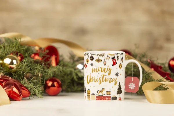 Primer plano de una taza de Navidad con colgar etiqueta de té vacío con alrededor de ramas de pino, adornos rojos y cinta de oro satinado sobre fondo de madera con efecto bokeh — Foto de Stock