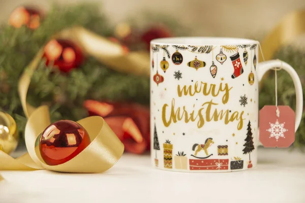 Close up of a Christmas mug with hanging empty tea label with around pine branches, red baubles and satin gold ribbon on wooden background with bokeh effect — Stock Photo, Image