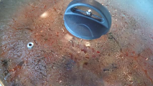 典型的なイタリアのオリーブとケッパーのパスタソースのクローズアップは 鍋で調理し 男の手は木製のスプーンでそれを混合し 調理蒸気で曇ったガラス蓋でそれをカバーします — ストック動画
