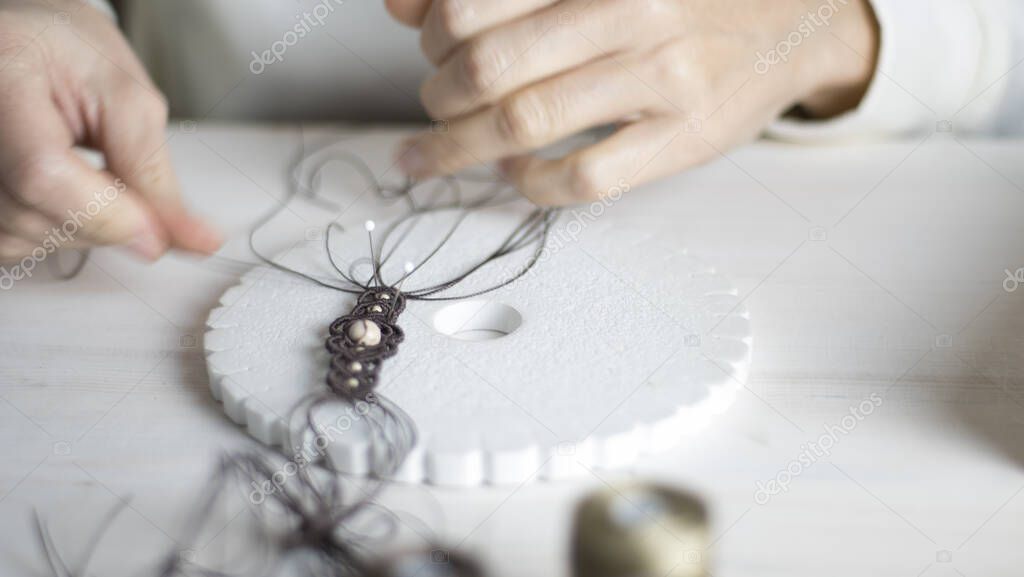 Lifestyle concept, work from home to reinvent your life: close-up of woman hands making macrame knotted jewelry with stone beads and tools on light wooden table