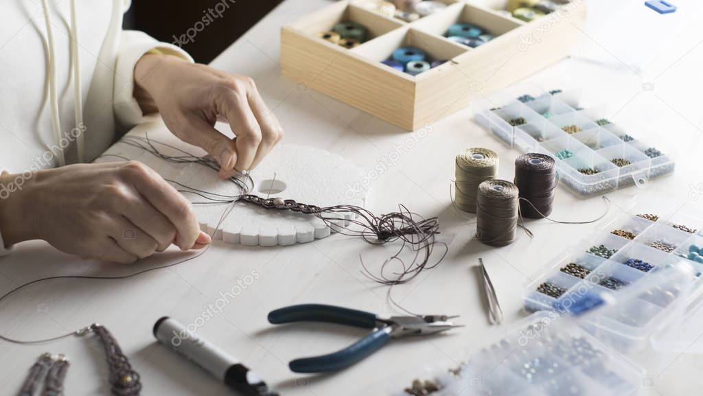 Lifestyle concept, work from home to reinvent your life: close-up of woman hands making macrame knotted jewelry with stone beads and tools on light wooden table