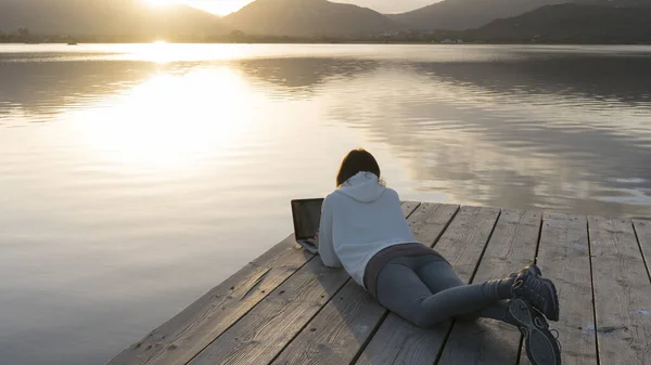 Internet freelance job choice concept: a young woman works on her laptop lying on a pier by a lake at sunset